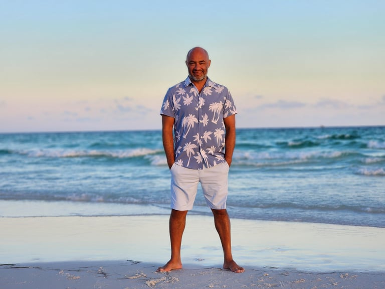 Bald man in blue floral shirt and white shorts standing on beach at sunset with ocean waves in background