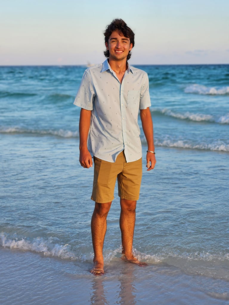 Man standing in shallow ocean water wearing a light blue shirt and tan shorts, smiling at the camera with a beach and blue sky in the background